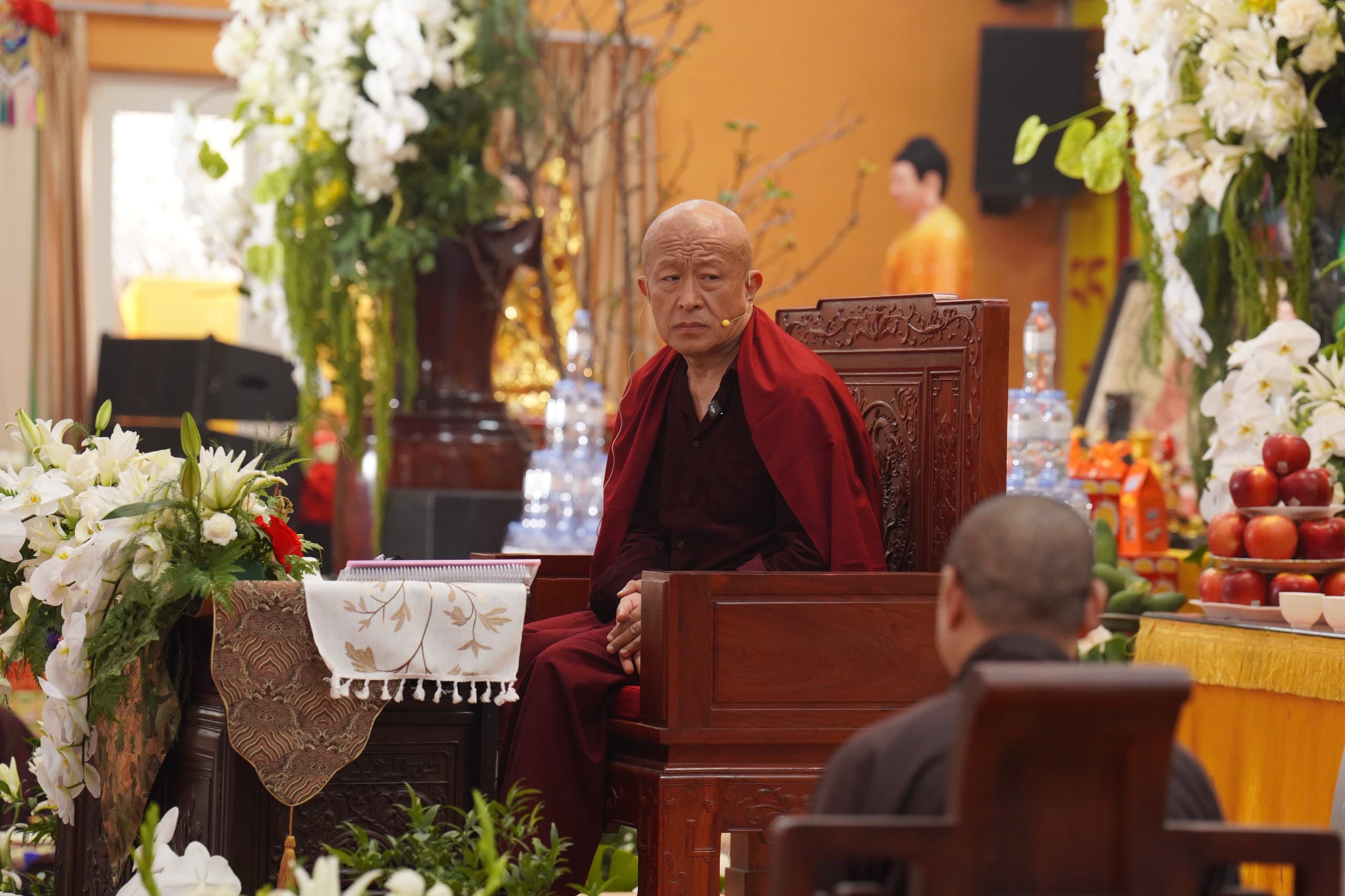 Vajradhara Dzongsar Jamyang Khyentse Rinpoche, in maroon robes, seated on a carved wooden teaching throne before altar offerings of white orchids, lilies and fruit.
