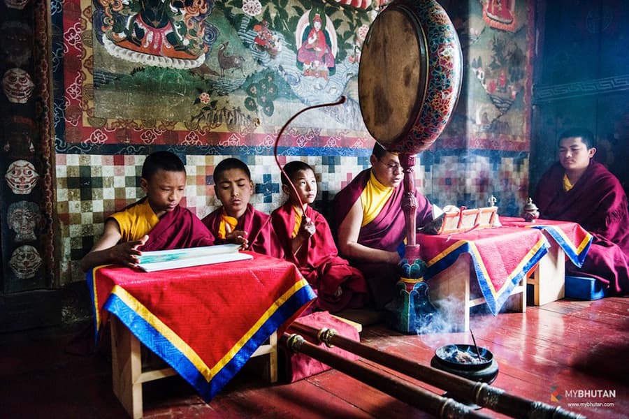 Young monks of the monastery seated at red-draped tables performing a ritual with a large ceremonial frame drum, against walls covered in traditional Buddhist murals.