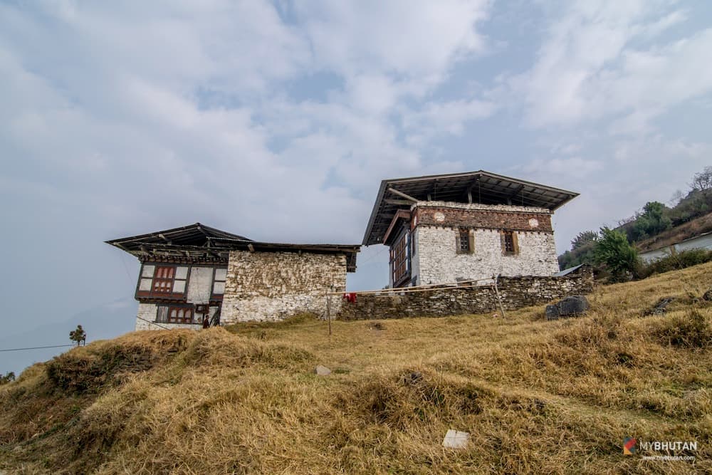 Two traditional whitewashed stone temple buildings with carved Bhutanese window frames on a grassy hillside beneath a soft, cloudy sky in the Mangde valley, Trongsa.